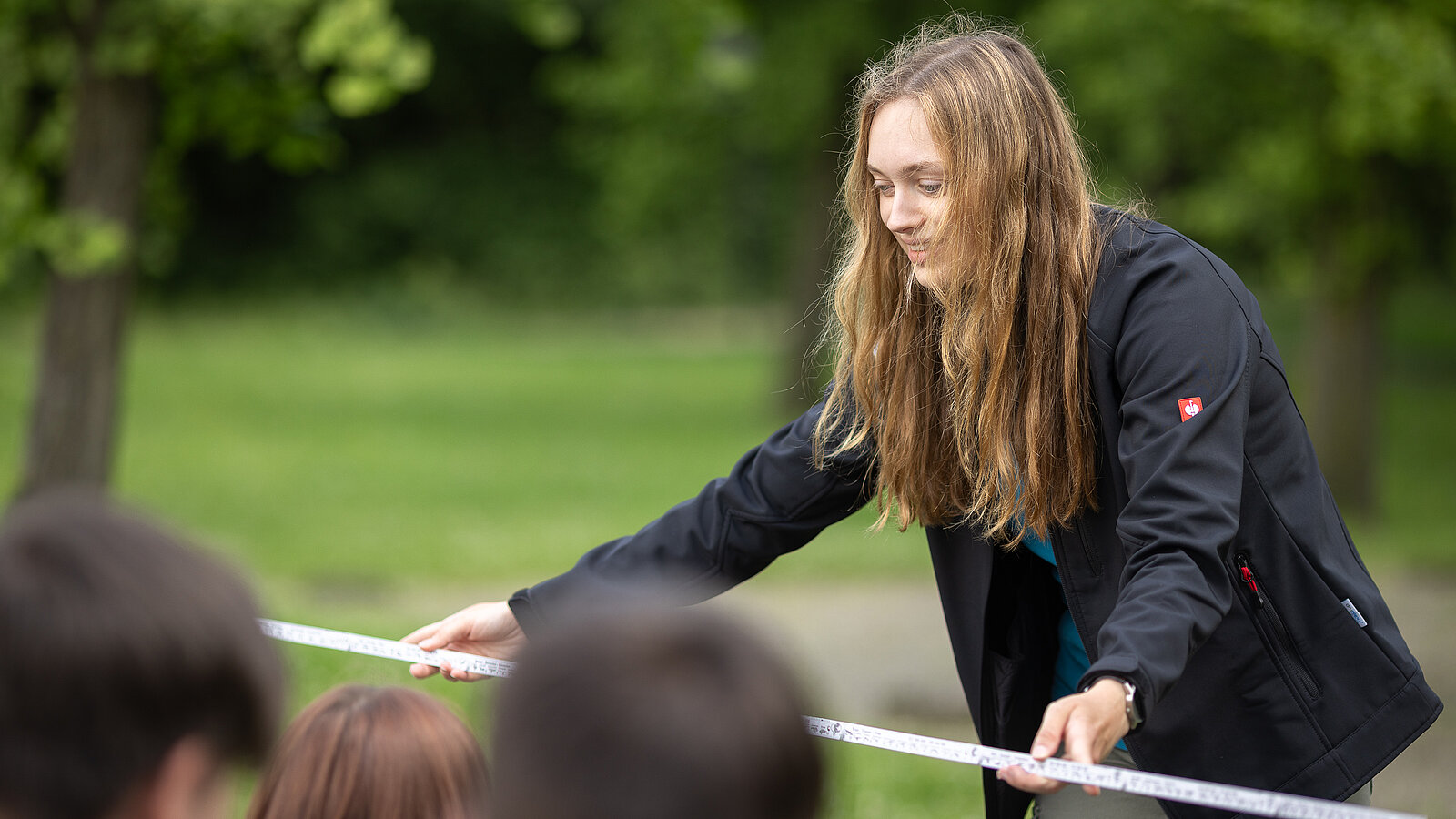 Eine Umweltbildnerin hält vor einer Schulgruppe einen Zollstock in der Hand