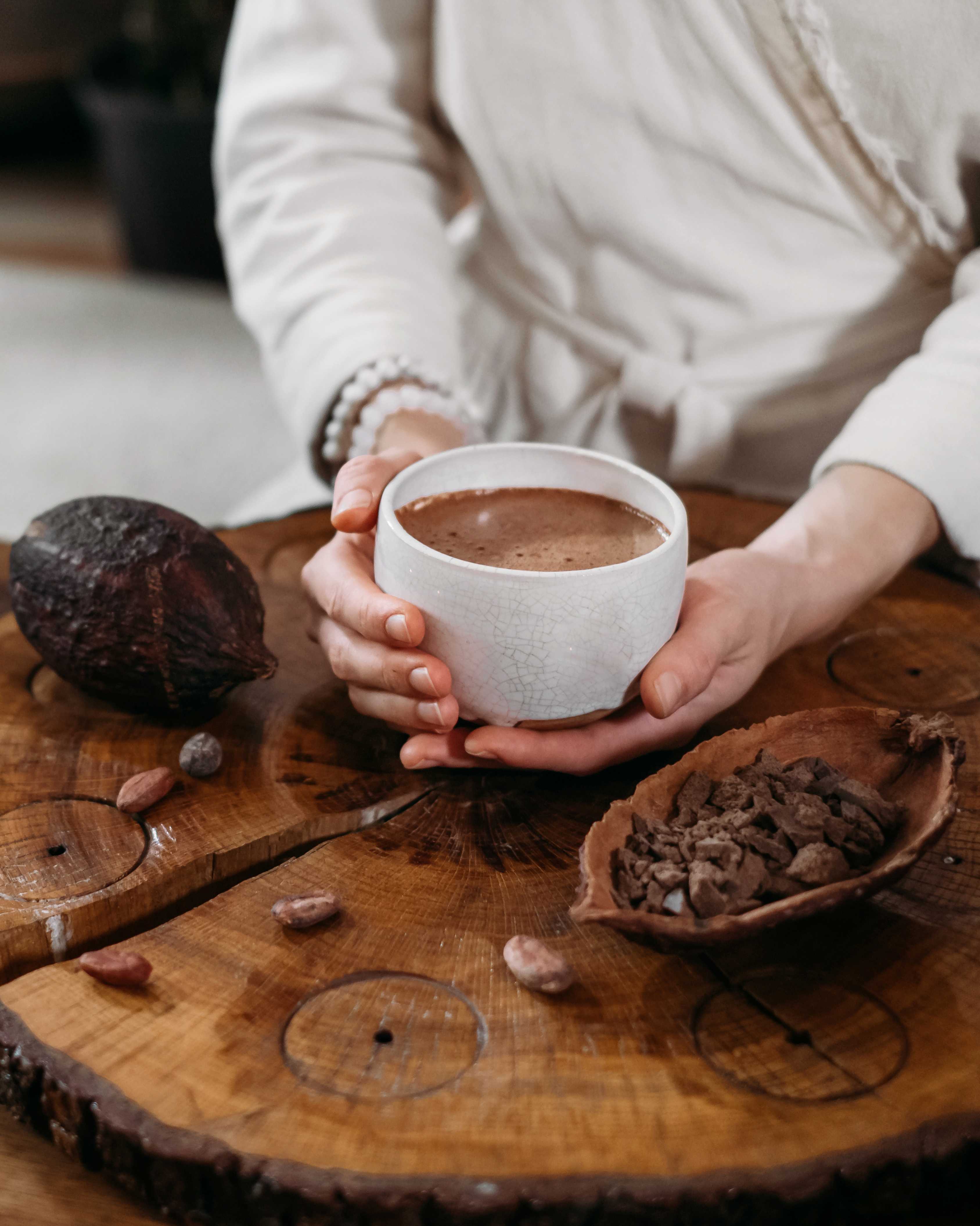 Eine Person hält eine Tasse mit Kakao in ihren Händen. Neben ihr liegen Kakeobohnen